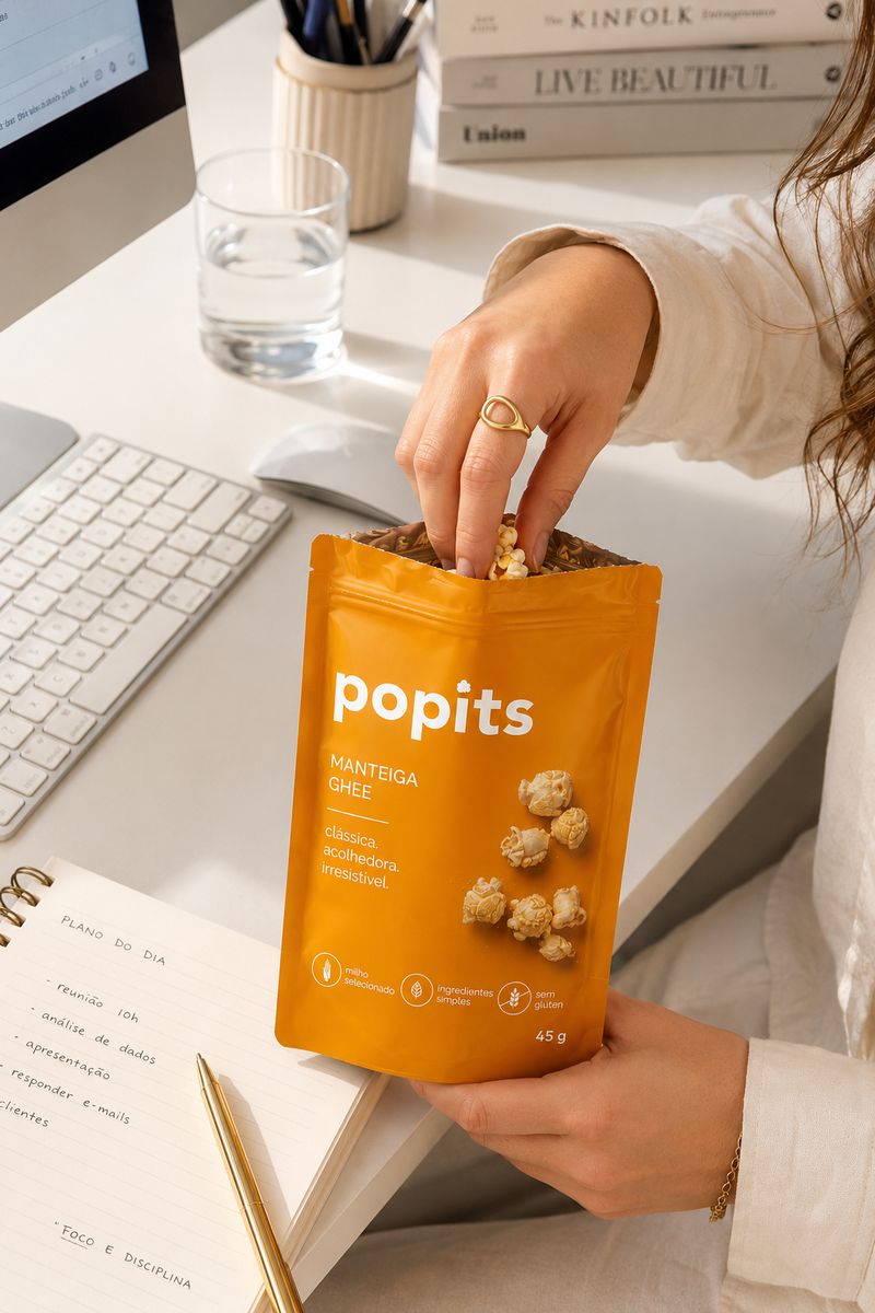 Mão pegando pipoca de um pacote Popits Manteiga Ghee na mesa de trabalho.